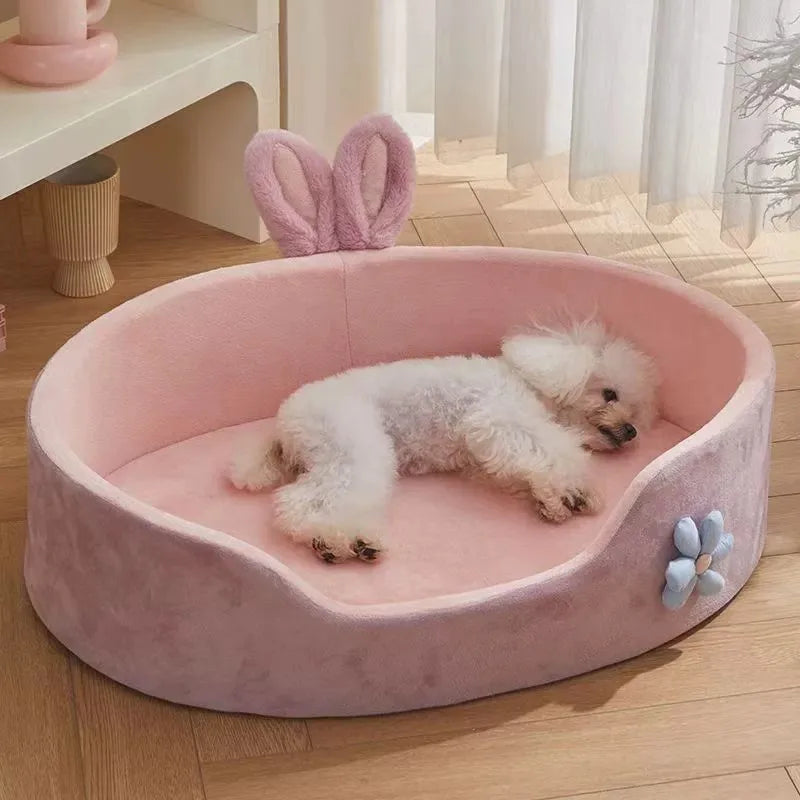 Small white dog lying on a pink pet bed with bunny ears in a room.