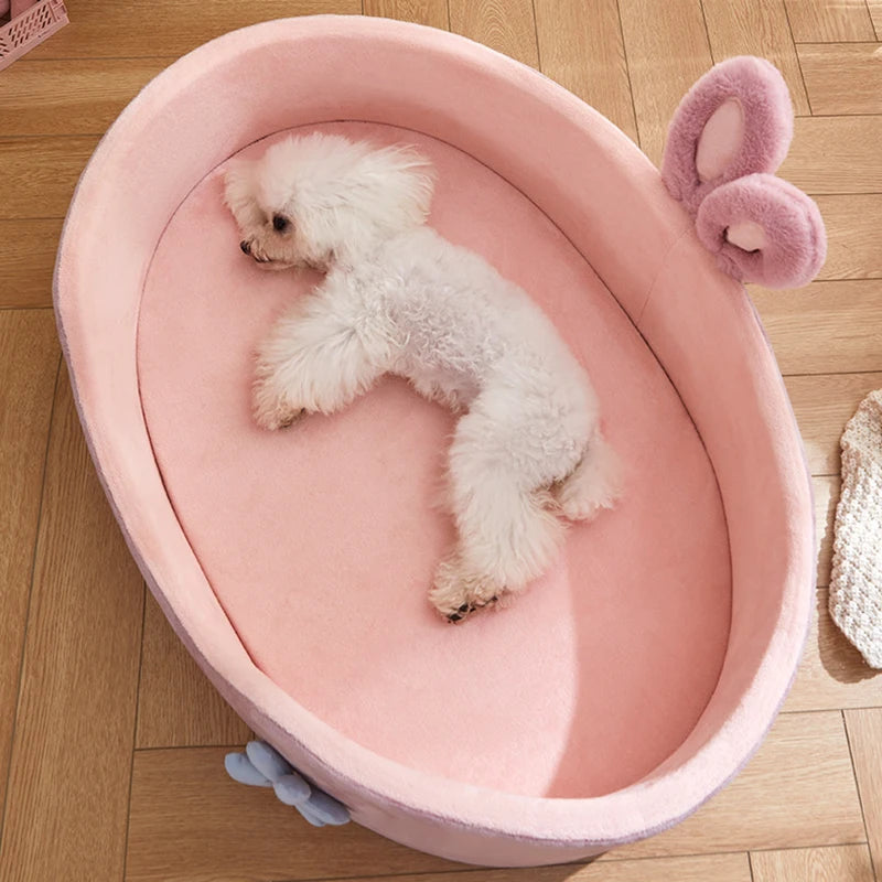 Small white dog lying in a pink heart-shaped pet bed on a wooden floor.